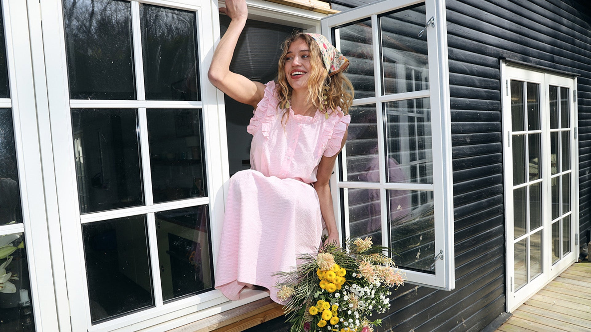 Woman in a light pink maxi dress sitting in an open window with a bouquet of wildflowers in hand outside a black wooden summer house.