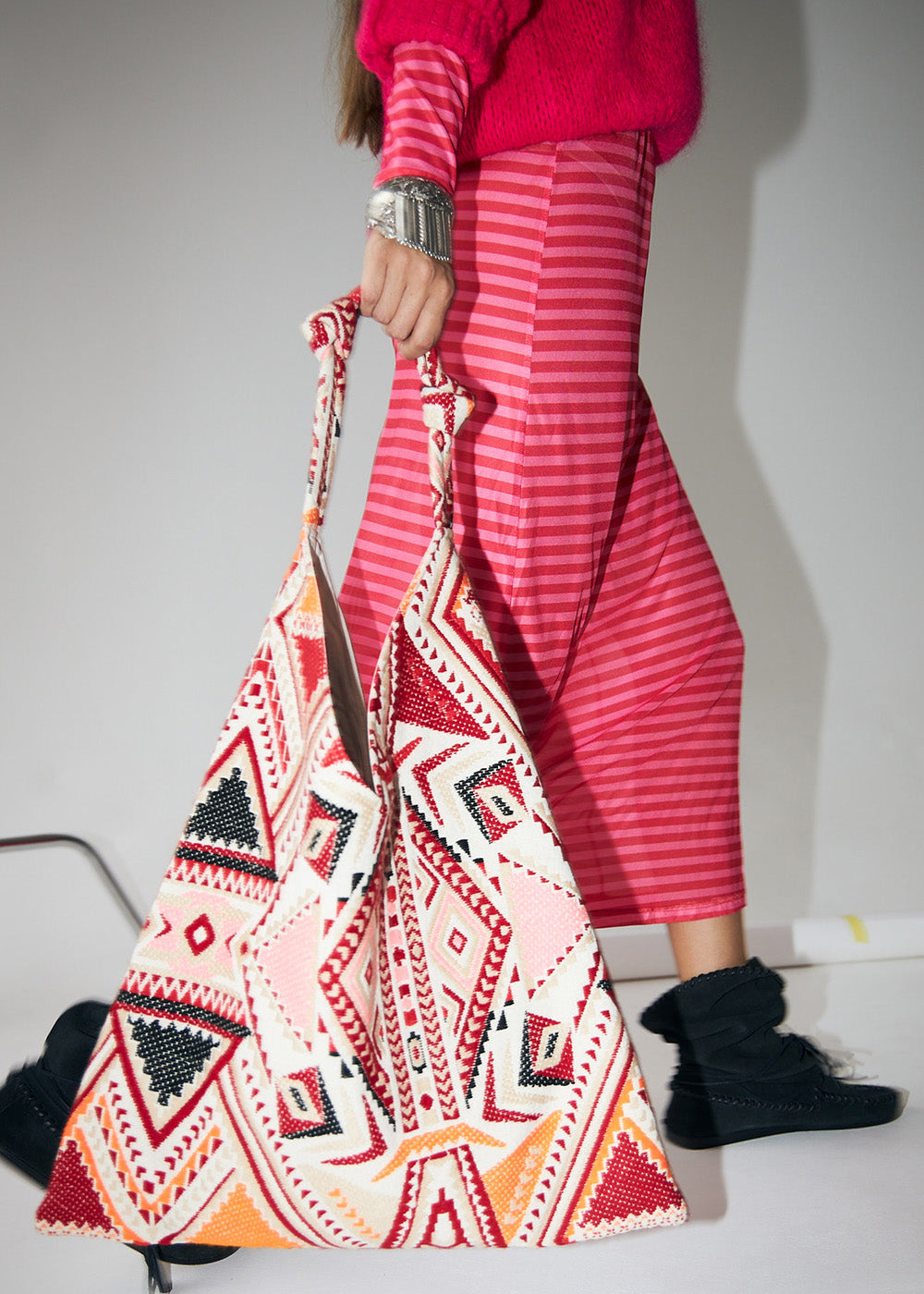 Model wearing a pink knit and striped skirt, carrying a large bag with a colourful geometric pattern in red, orange, and pink tones from Lollys Laundry.