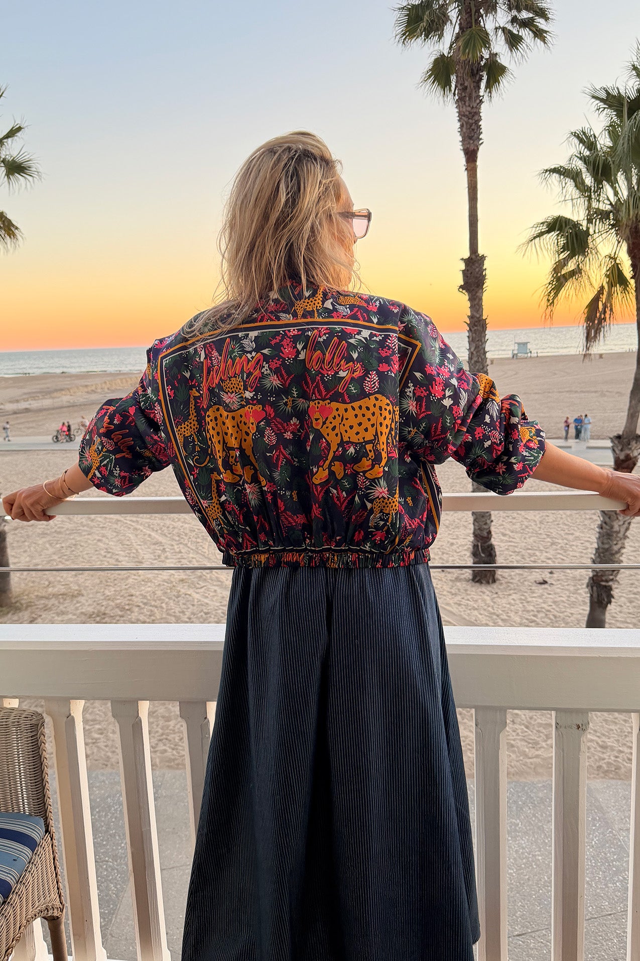 Woman standing on a balcony at sunset overlooking the beach, wearing a colorful Lollys Laundry jacket with leopard motifs and “feeling Lollys” lettering in the print, paired with a dark blue skirt with fine white pinstripes.