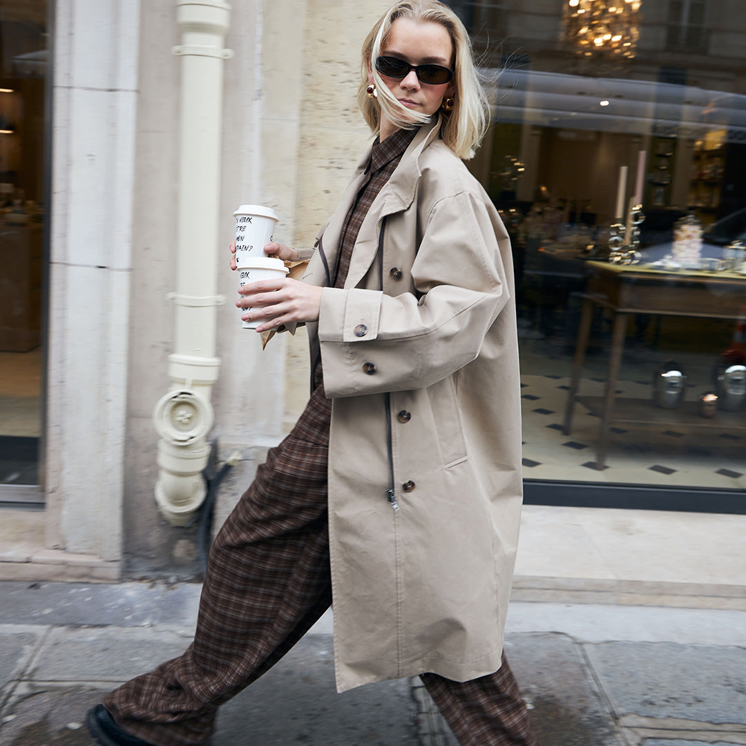 Woman in a beige trenchcoat holding a coffee cup on a city street