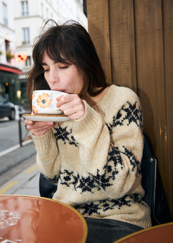 A woman with dark hair sits at an outdoor cafe table, holding a white ceramic coffee cup with a floral pattern and saucer. She is wearing a cream-colored knit sweater with a black nordic pattern, leaning against a wooden wall.