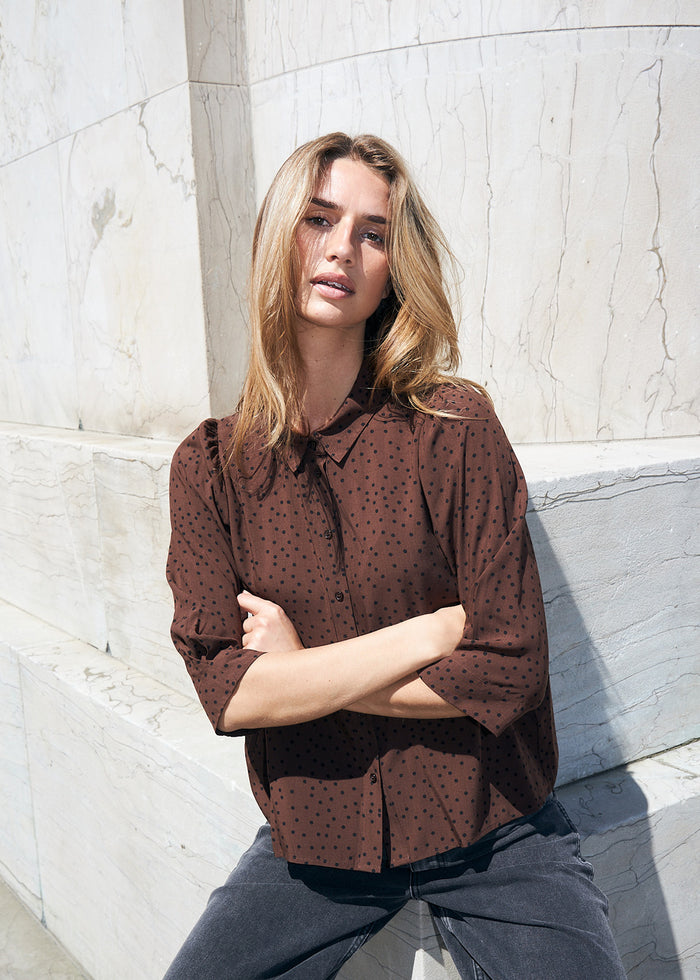 Woman wearing a brown polka dot shirt with a collar and three-quarter sleeves, styled with dark grey jeans.