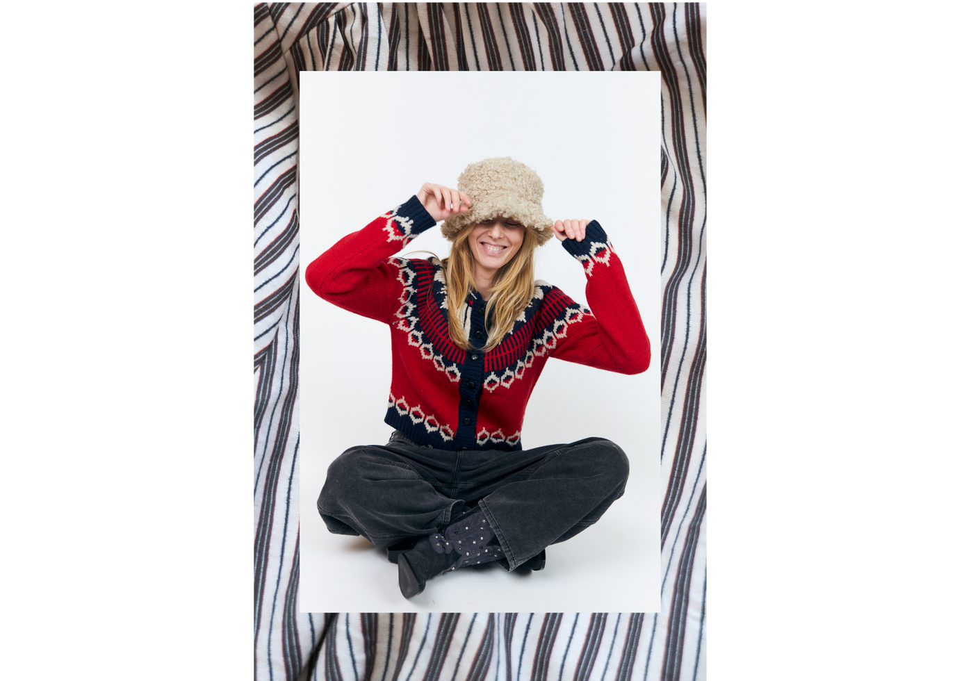 Woman wearing a red cardigan with a Nordic pattern, dark jeans and a beige teddy bucket hat. A colourful autumn look with soft knitwear and warm accessories.