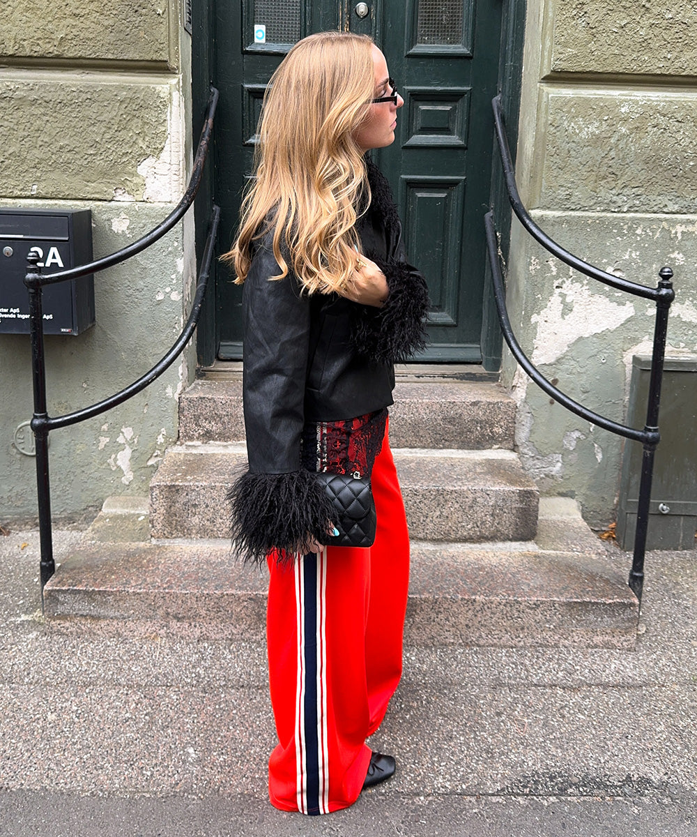 Woman wearing a black jacket with fluffy sleeve details and red trousers with side stripes, styled with sunglasses and a black bag in front of a green door.