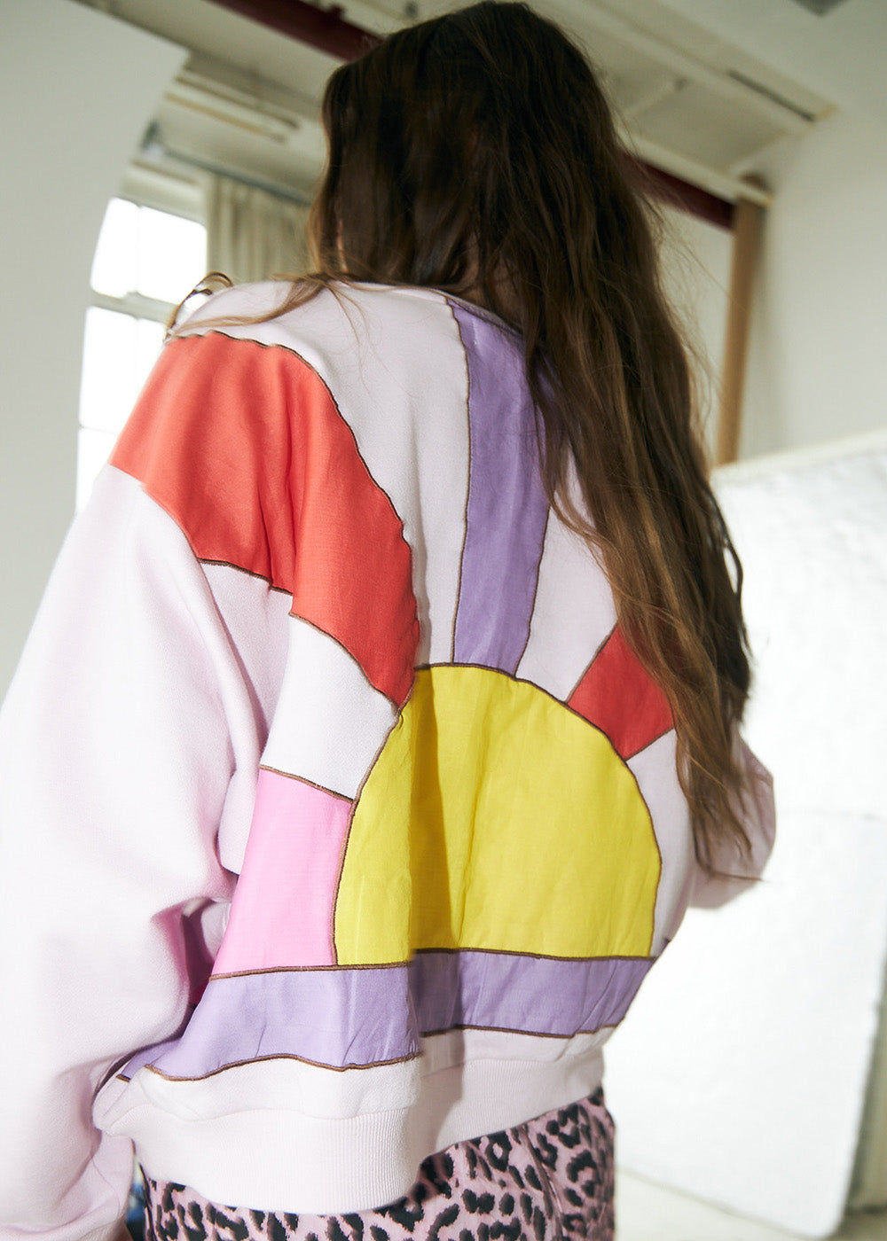 Model wearing a light lilac sweatshirt with a sunrise motif in yellow, purple, pink, and orange tones on the back, styled with pink leopard-print trousers from Lollys Laundry.