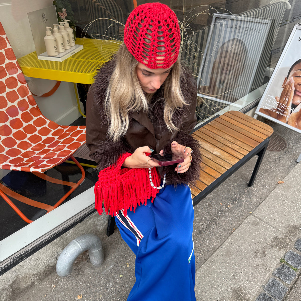 A woman sits on a wooden bench outside a shop, wearing a bright red crocheted beanie, a brown faux-fur jacket, a red fringed bag, a blue skirt, and leopard-print shoes while looking at her phone.