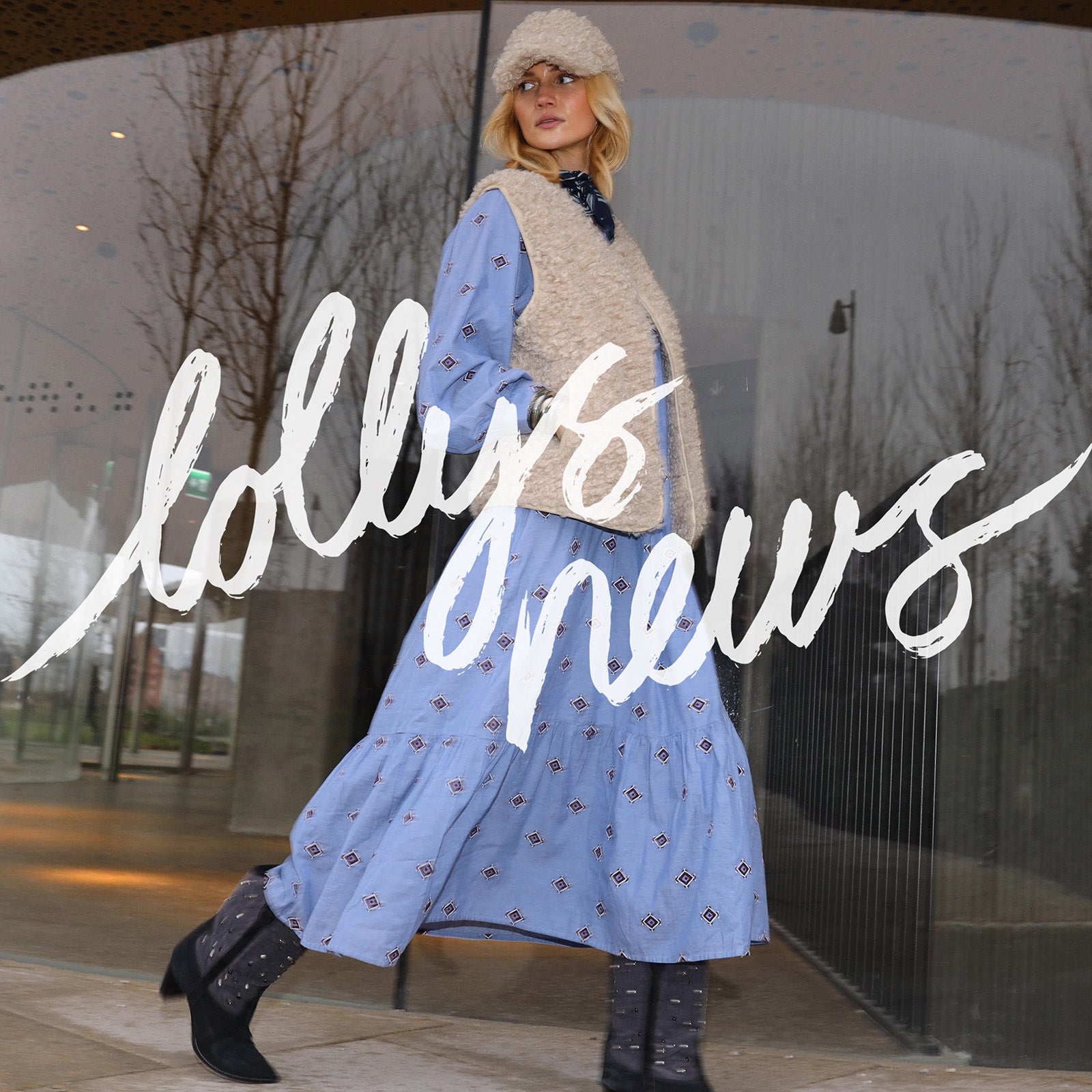 Woman in a blue dress and beige teddyvest standing in front of a glass building with 'lollys news' branding.