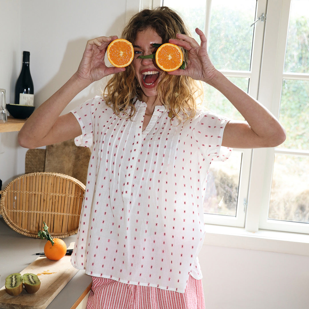 Smiling woman indoors holding two orange halves in front of her eyes, wearing a light blouse with small light pink dots and striped bottoms in red, creating a playful, summery mood.