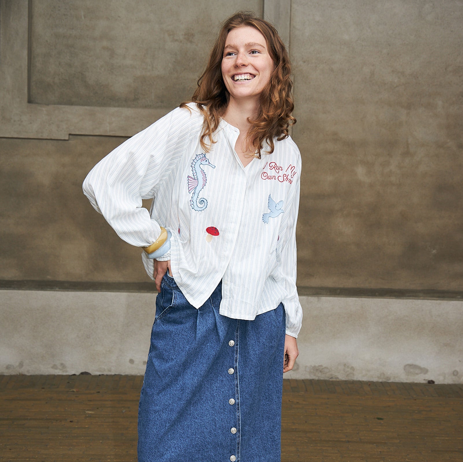 A woman wearing a white long-sleeve button-up shirt with colorful embroidered seahorse and butterfly motifs, paired with a blue denim skirt, standing against a textured concrete wall.
