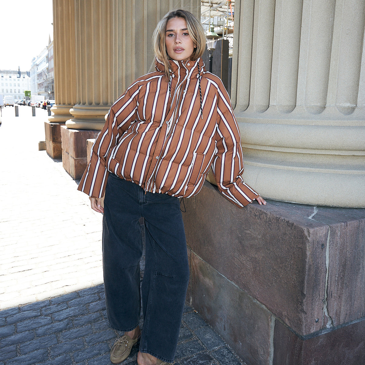 Woman standing in front of large stone columns, wearing a brown hooded jacket with white and dark stripes, wide sleeves and dark jeans.