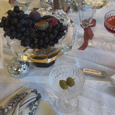 Festively decorated Christmas table with grapes, fruit, crystal glasses, disco balls and decorative details on a white tablecloth.