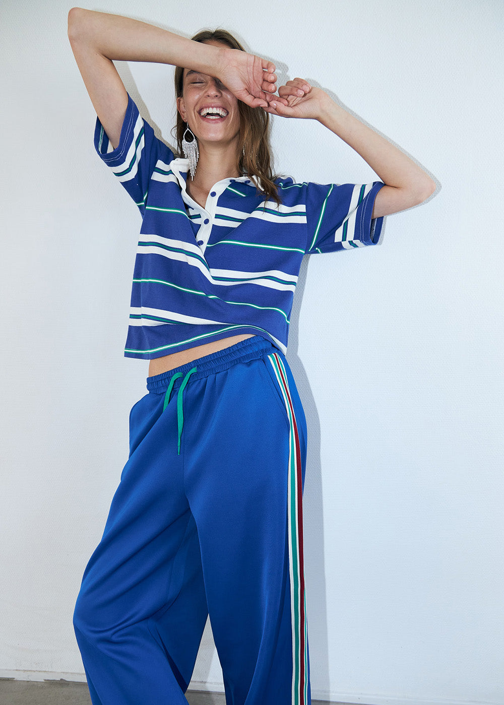 Smiling woman wearing a blue striped polo top styled with matching blue pants with side stripes, photographed in a bright studio.